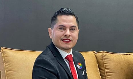 Latino man sitting on brown couch smiling, wearing red lipstick, red tie, black suit jacket, white shirt, rainbow pin.