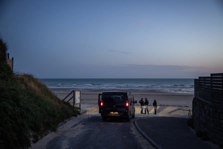 Police officers stand around their van in the early morning light at the beach as the sun comes up over the sea; the van’s taillights are illuminated.