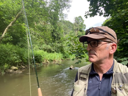 Middle-aged man with baseball cap and glasses holding a fishing rod by a river