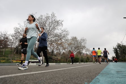 Men and women jog on a track through a park. The woman at the front of the picture wears a T-shirt and leggings and has her head uncovered.