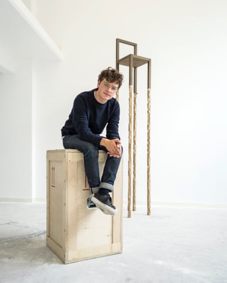 A young man sits on a wooden packing crate in front of a simple chair on stilt-like legs