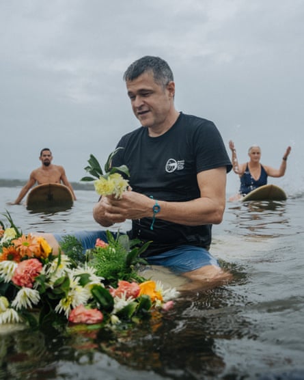 A man holding a flower straddles a surfboard the top of which is covered in flowers