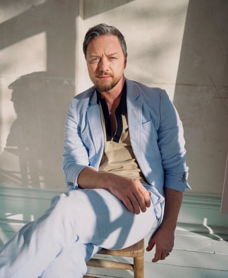 James McAvoy sitting on a small wooden chair, wearing pale blue trousers and jacket, over a brown shirt and fawn v-neck jumper, with shadows