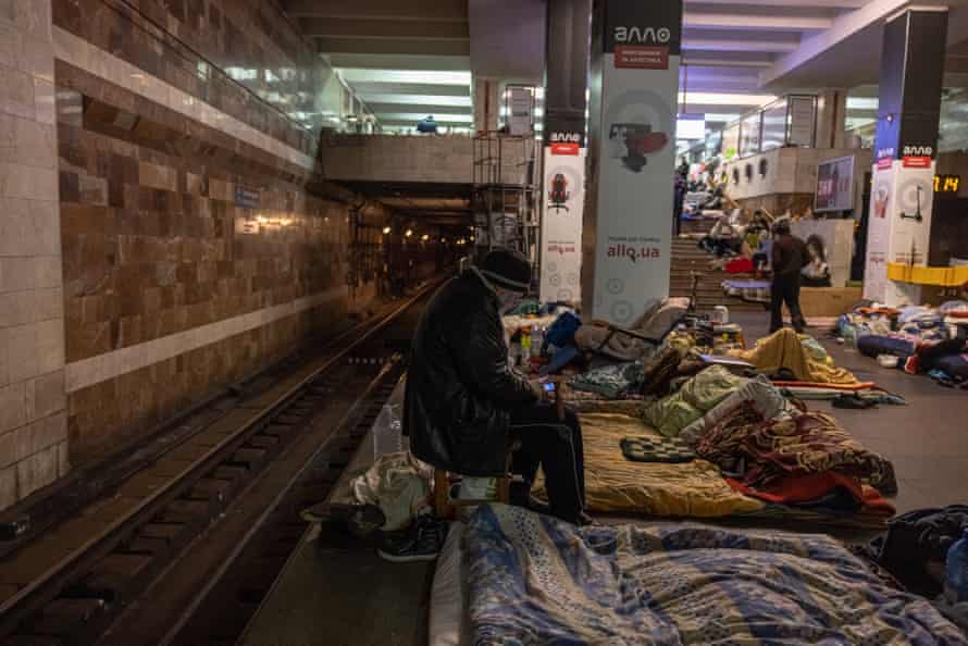 A man sits inside a subway station with a bed close to the railway tracks.