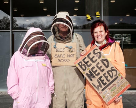 Three people in beekeeper suits, one of them holding a sign reading ‘Bees need weeds’