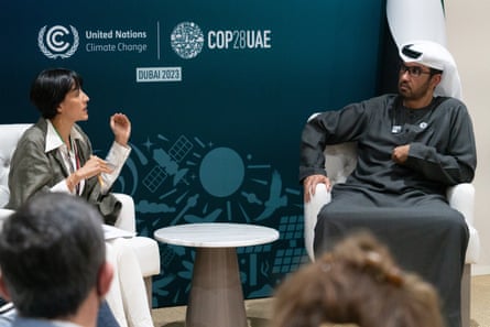 A woman and a man conversing as they sit on armchairs in front of a teal backdrop with the Cop28UAE logo on it.