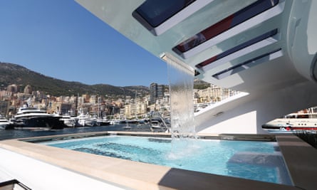 The rear deck of a superyacht with a striking overhanging glazed canopy, through which a wide stream of water is falling into a small swimming pool below