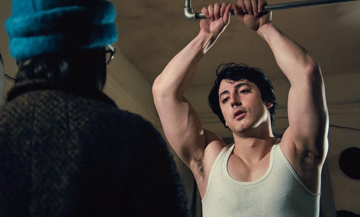 Anthony Ippolito as Sylvester Stallone is wearing a white vest and holding on to a bar above his head while someone wearing a thick coat and hat stand with his/her back to the camera, facing him