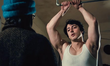 Anthony Ippolito as Sylvester Stallone is wearing a white vest and holding on to a bar above his head while someone wearing a thick coat and hat stand with his/her back to the camera, facing him