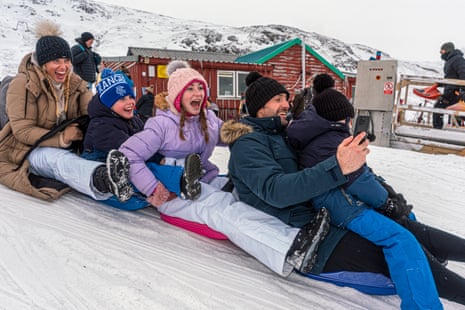 Two adults and three children looking happy on sled with man taking photo