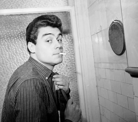 The young Ian McShane adjusts his tie and smokes a cigarette in front of a wall-mounted mirror