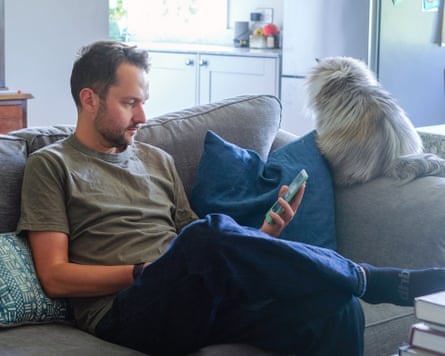 The writer is sitting on a sofa, looking at an e-reader with his cat on the back of the sofa