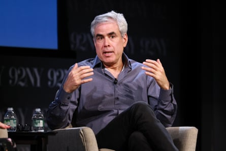 He sits in an upholstered chair on a stage with both arms raised in front of his chest. He is mid-conversation