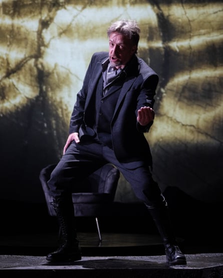 a man in a dark suit (Julian Close playing Hagen) stoops while singing, against a foreboding stage backdrop in a scene from Götterdämmerung at Longborough Festival Opera.