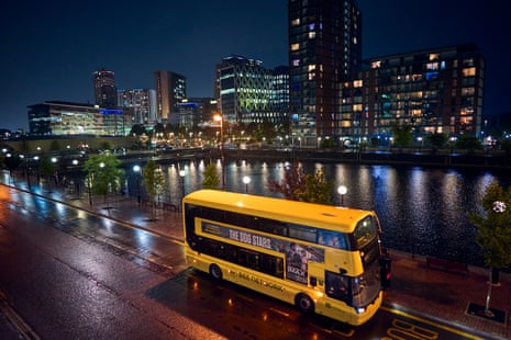 Buildings light up Salford Quays in the evening as a yellow bus travels past.