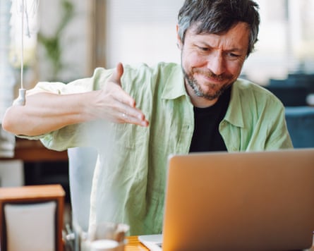 A mature man in a green shirt waves his hand and gestures while working on a laptop.