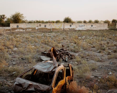 A scruffy field scattered with debris and a ruined car in the foreground. In the distance, a wall with IS logos on