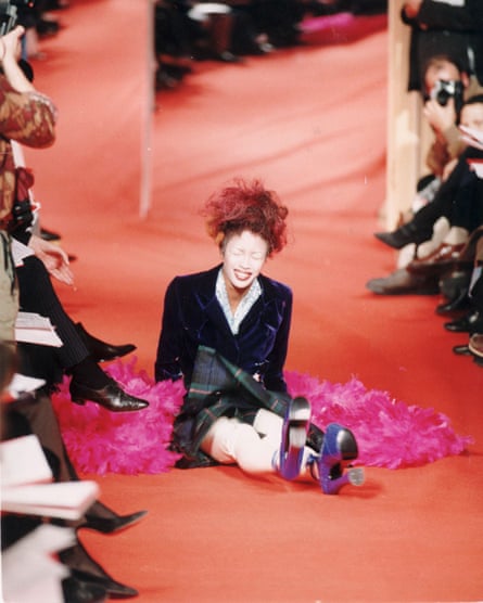 Naomi Campbell sits on an orange catwalk floor laughing after falling over in very high blue platforms