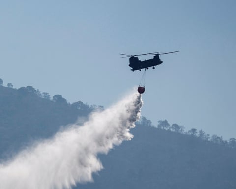 A helicopter silhouetted against a blue sky releases water over a forested hillside