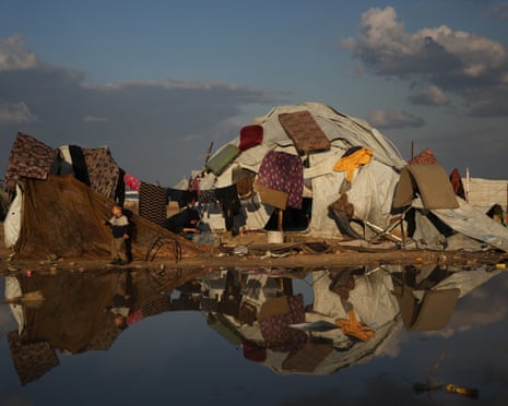 A Palestinian boy stands next to his mother washing clothes as mattresses hang to dry over their tent in a makeshift camp