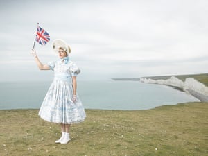Perry, deadpan in doll-like dress and lace bonnet, stands near the edge of the grassy cliff overlooking the sea, holding up a small union flag