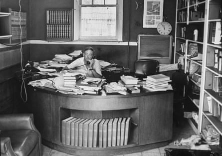 A man speaks on the phone at his curved desk that is cluttered with papers and books