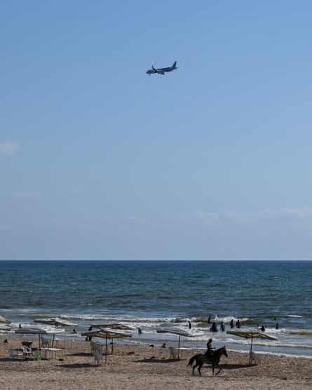 A plane flies over a beach and waves, with people relaxing under parasols and a man riding a horse