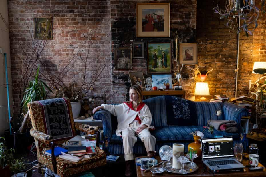 A person wearing white and red sits on a blue couch in a living room with brick walls and framed artwork