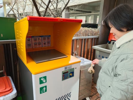 A woman approaches a bin with a digital keypad on the side with a small bag of food waste