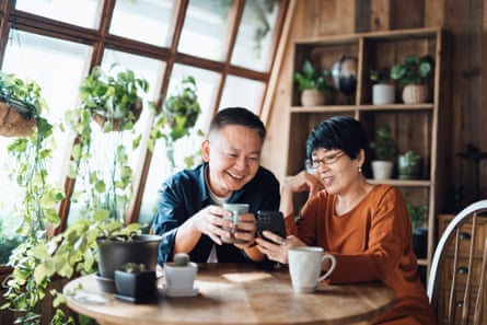 Happy senior Asian couple sitting at a wooden table looking at a phones as if they are video-chatting; he has short dark hair and holds a mug, while she has neat black hair, glasses and a dark orange top; there are wooden shelves of pottery behind them and large sloping glass windows with trailing houseplants in hanging baskets.