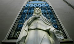 Stone statue of young woman in hooded robe with palm out,  beneath stained glass window outside building.
