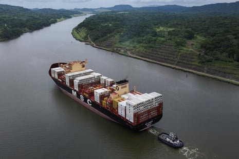 A container ship transits the Panama canal through a forested waterway, followed by a tugboat
