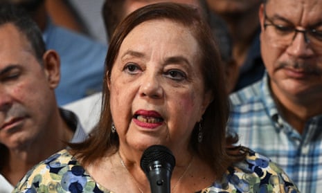 Older Latina woman with dyed brown-red hair, patterned, dress, red lipstick, speaks into a microphone surrounded by people.