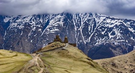 The Gergeti Trinity church under Mount Kazbek in Georgia. Church perched on green hilltop with vast dramatic mountains in the background sprinkled with snow