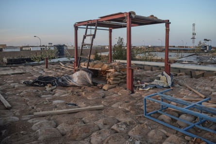 A general view of damaged infrastructure at the army camp: there are skeletal iron frames of structures and piles of sandbags plus strewn debris. There are low-rise buildings in the background in the desert landscape.