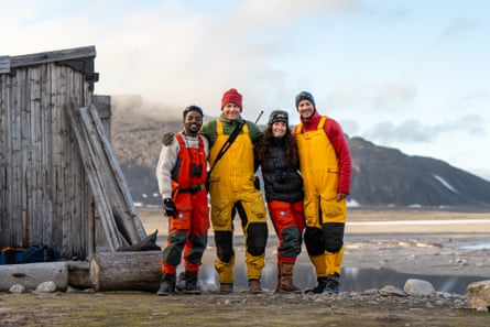 Group shot of four people in orange and yellow waterproof bib suits and hats next to a wooden hut by the sea