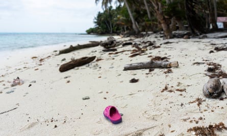 A dark pink child’s Croc sandal lies on a long white sand beach strewn with driftwood, with blue water to the left and palm trees to the right.