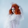 Chappell Roan with voluminous orange hair and colourful makeup wears a white textured, fringed garment