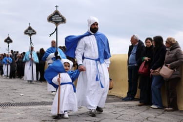 Image 20: A man holds the hand of a child as they join a procession. They are dressed in costume