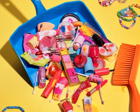 A blue dustpan full of garishly coloured makeup products, next to an orange brush on a yellow floor.