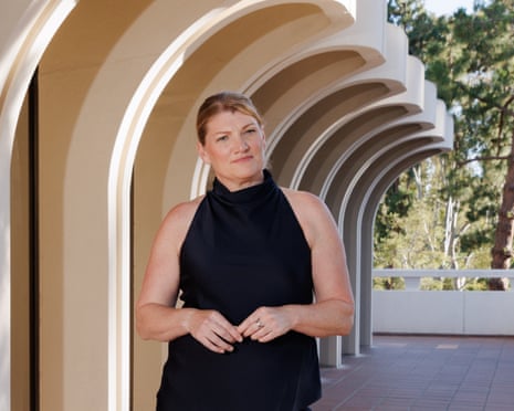 Wearing a black halterneck top, she poses for a picture with her arms in front of her torso. She stands on a wraparound balcony outside a modern building, with trees in the background
