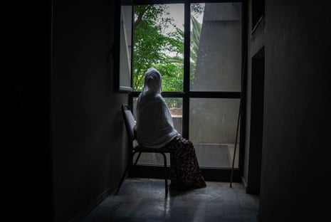 A woman sits in front of a window with her back to the camera, her head covered.