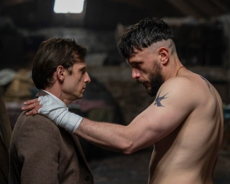 A shirtless man with tattoos puts his arm on the shoulder of a shorter man wearing a suit as they look at each other