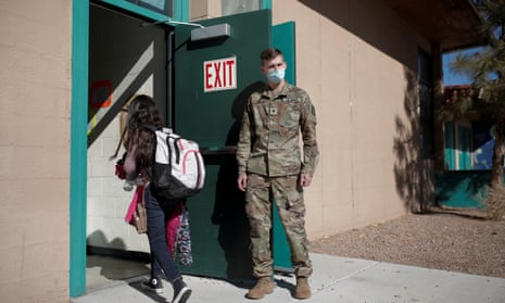 National guard Specialist Austin Alt, wearing an olive green camouflage uniform, holds open a classroom door as a young girl wearing a backpack walks in.