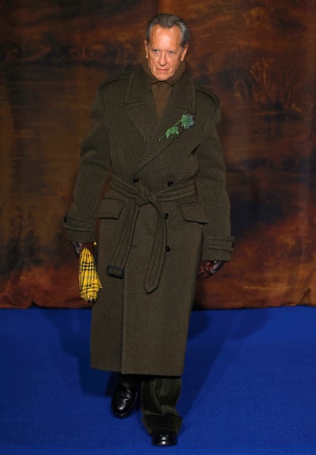 Richard E Grant on stage wearing a long brown tweed coat over a brown jumper, black trousers and black shoes