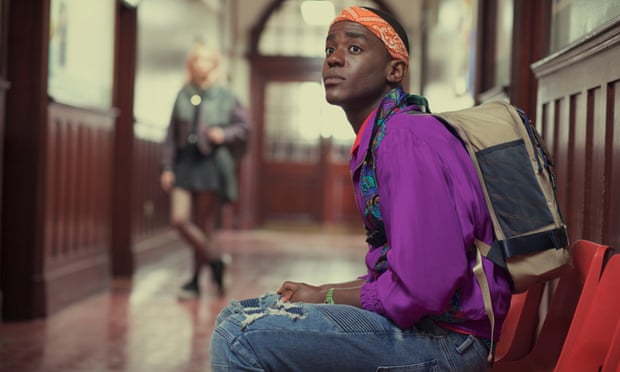 Ncuti Gatwa as Eric Effiong, wearing a purple top and denim dungarees and sitting in a chair in a school corridor, staring and looking slightly anxious, in a scene from Sex Education.