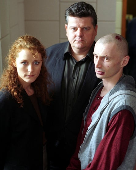 A red-haired woman in a black suit stands next to a dark-haired man in a black suit and a shaven-headed man with cuts to his face and head.