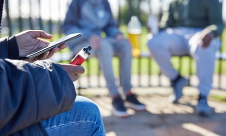 Three teenagers with mobile phones, vaping and drinking in a park.
