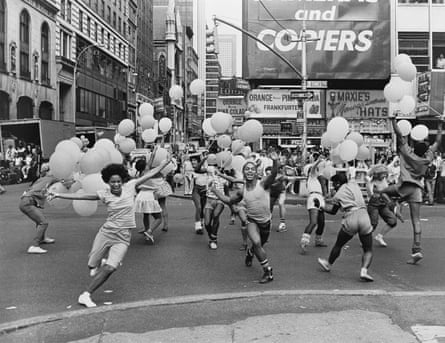 Several lines of dancers on a busy New York City street strut around and wave balloons