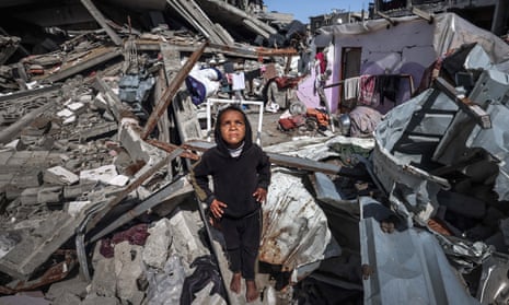 A young girl looks upwards, standing on a pile of rubble. Everything around her is destroyed.